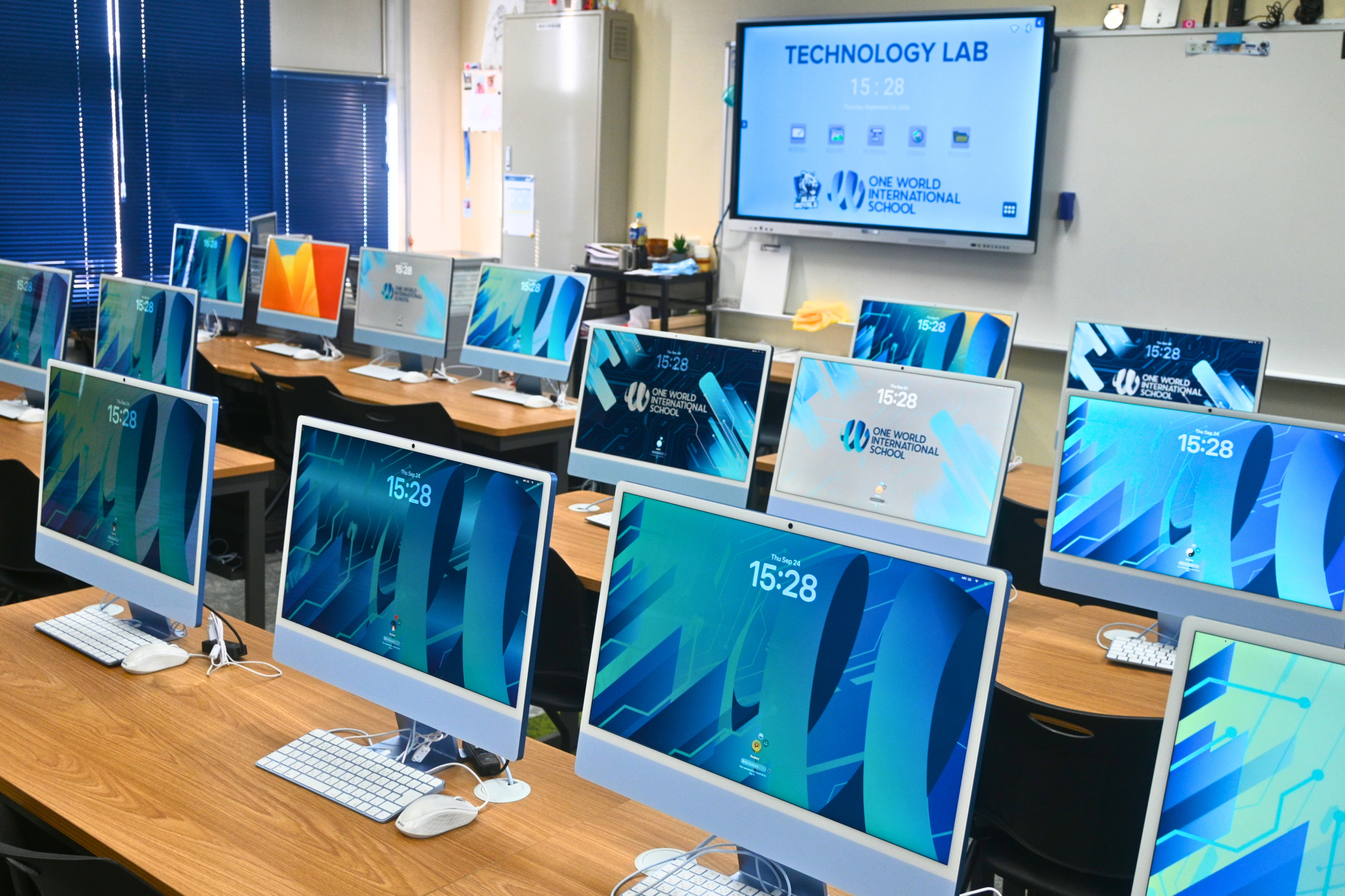 Information Technology (IT) Lab at One World International School Osaka, showing OWIS branded screensavers and desktop backgrounds on Apple PC screens in an IT classroom